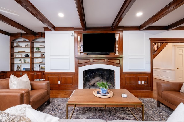 living area featuring wood finished floors, beamed ceiling, a large fireplace, a decorative wall, and built in shelves