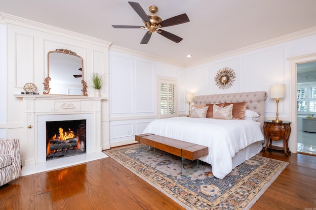 bedroom with a decorative wall, crown molding, wood finished floors, a lit fireplace, and multiple windows