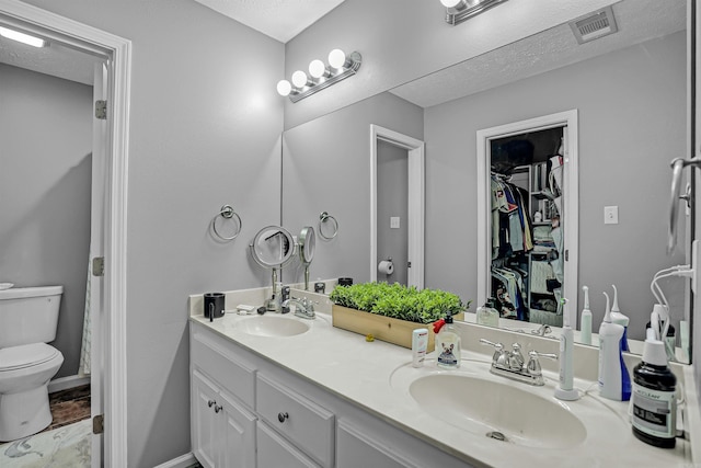bathroom with toilet, double vanity, a walk in closet, a textured ceiling, and baseboards