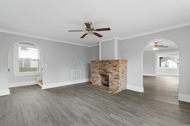 unfurnished living room with ceiling fan, arched walkways, a brick fireplace, wood finished floors, and ornamental molding