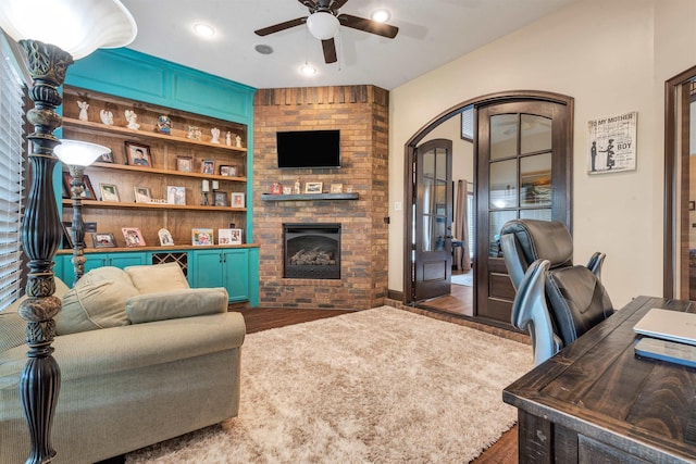 living area featuring a brick fireplace, ceiling fan, dark wood finished floors, arched walkways, and built in shelves
