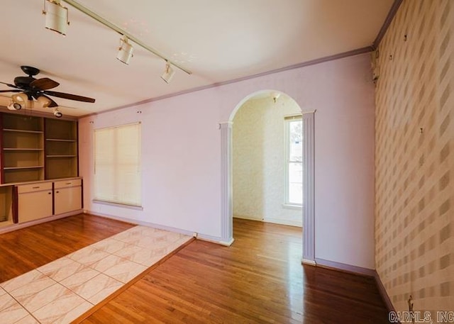 unfurnished living room with wallpapered walls, track lighting, arched walkways, wood finished floors, and ornamental molding