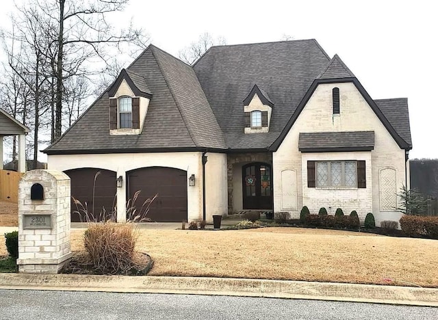 french country style house with roof with shingles, a garage, driveway, and brick siding