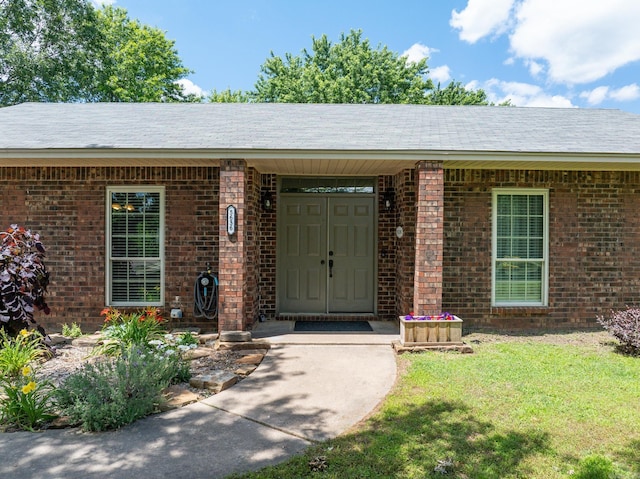 view of exterior entry featuring a lawn, brick siding, and roof with shingles