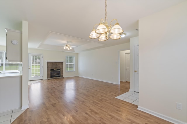 unfurnished living room featuring a ceiling fan, a tray ceiling, light wood finished floors, a brick fireplace, and a chandelier