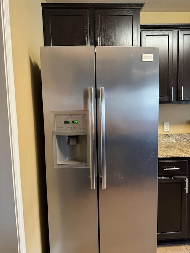 kitchen view of stainless steel refrigerator with ice dispenser, dark brown cabinets, and light stone countertops