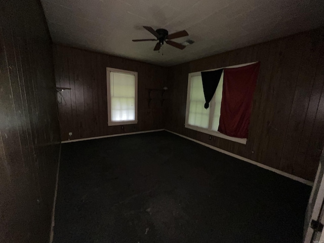 empty room with wood walls, a ceiling fan, healthy amount of natural light, and dark colored carpet
