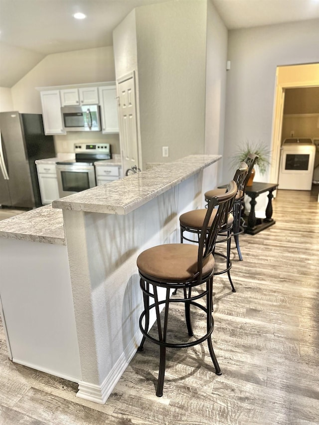 kitchen with appliances with stainless steel finishes, white cabinets, light wood-type flooring, a kitchen breakfast bar, and vaulted ceiling
