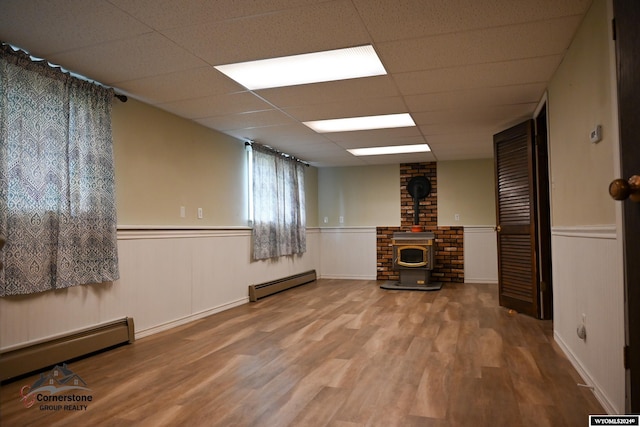 basement featuring brick wall, a baseboard radiator, a wood stove, a drop ceiling, and hardwood / wood-style floors