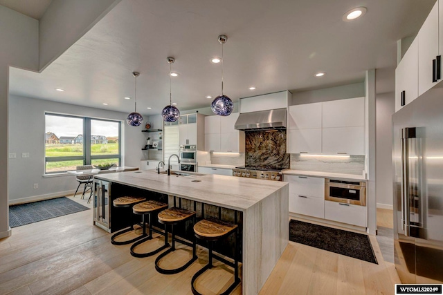 kitchen featuring decorative light fixtures, backsplash, a center island with sink, white cabinetry, and wall chimney exhaust hood