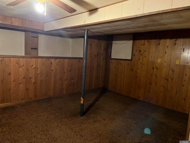 basement featuring ceiling fan, wood walls, and dark carpet