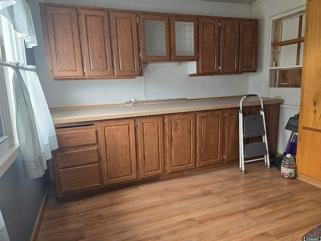 kitchen featuring brown cabinetry, glass insert cabinets, light wood-style floors, and tile counters