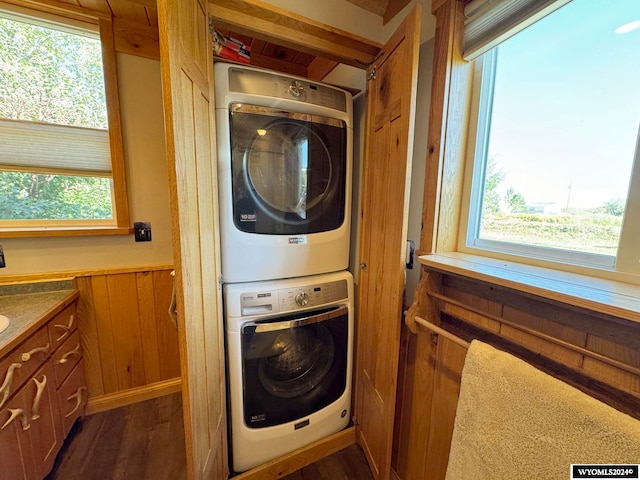 clothes washing area featuring stacked washer / dryer, a healthy amount of sunlight, wooden walls, and dark wood-type flooring