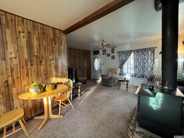 carpeted dining space with beam ceiling, a wood stove, ceiling fan, and wooden walls