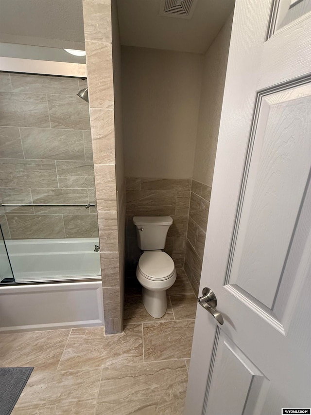 bathroom with toilet, combined bath / shower with glass door, and tile walls