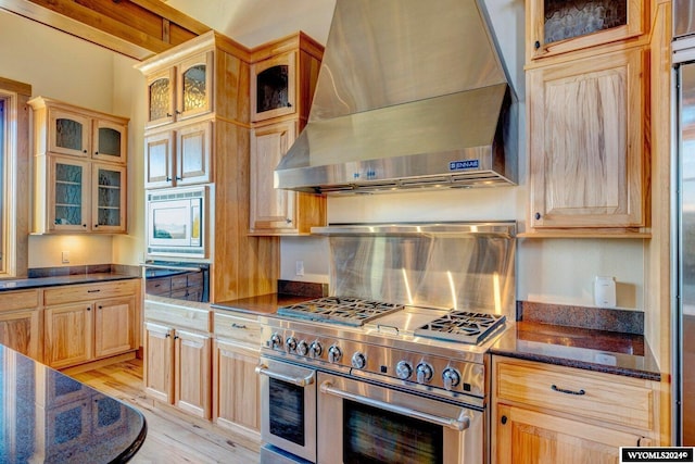 kitchen featuring light brown cabinets, wall chimney range hood, dark stone countertops, light wood-type flooring, and appliances with stainless steel finishes