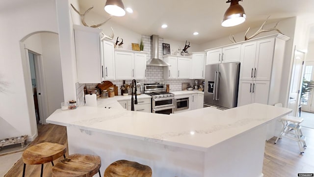 kitchen featuring kitchen peninsula, a kitchen breakfast bar, wall chimney exhaust hood, stainless steel appliances, and decorative light fixtures