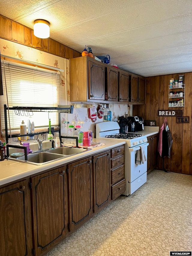 kitchen with dark brown cabinets, a textured ceiling, wooden walls, sink, and white range with gas stovetop