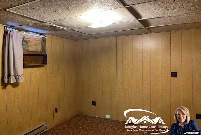 basement featuring a baseboard heating unit, a paneled ceiling, carpet flooring, and wooden walls