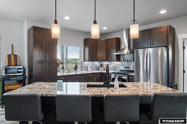 kitchen with stainless steel appliances, a sink, an island with sink, dark brown cabinets, and wall chimney exhaust hood