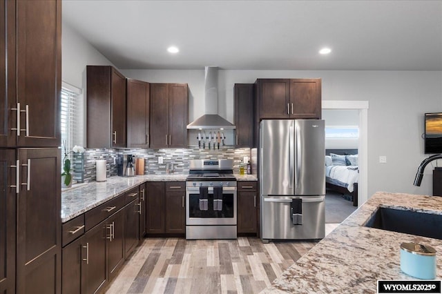 kitchen featuring light stone counters, decorative backsplash, appliances with stainless steel finishes, a sink, and wall chimney exhaust hood