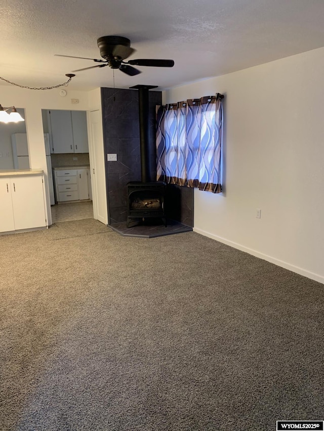 unfurnished living room with a textured ceiling, ceiling fan, carpet flooring, baseboards, and a wood stove