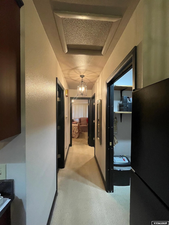 corridor featuring a notable chandelier, a textured ceiling, baseboards, and light colored carpet