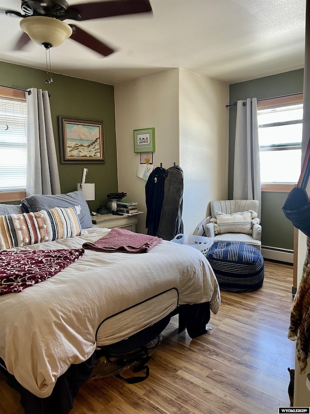 bedroom with a ceiling fan, multiple windows, baseboard heating, and wood finished floors