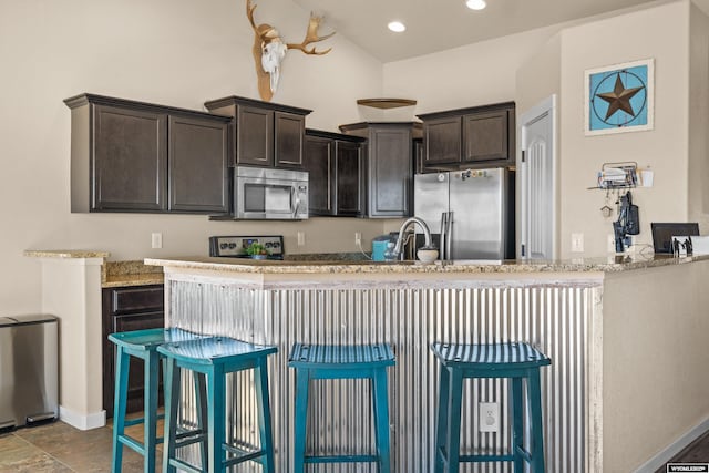 kitchen with recessed lighting, stainless steel appliances, dark brown cabinets, light stone countertops, and a kitchen bar