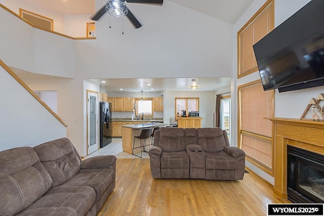 living room featuring light wood-style flooring, a high ceiling, ceiling fan, and a glass covered fireplace