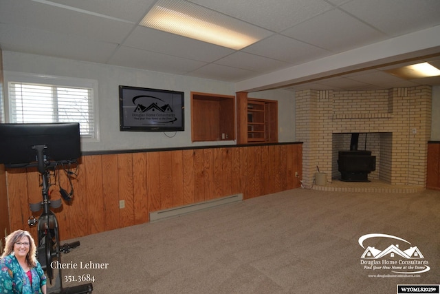 unfurnished living room with a baseboard radiator, a paneled ceiling, a wainscoted wall, wood walls, and a wood stove