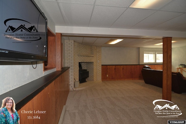 carpeted living room with a paneled ceiling, a wainscoted wall, a wood stove, and wood walls