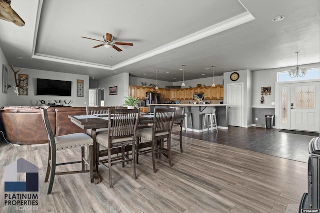 dining space with ceiling fan with notable chandelier, wood finished floors, a raised ceiling, and baseboards