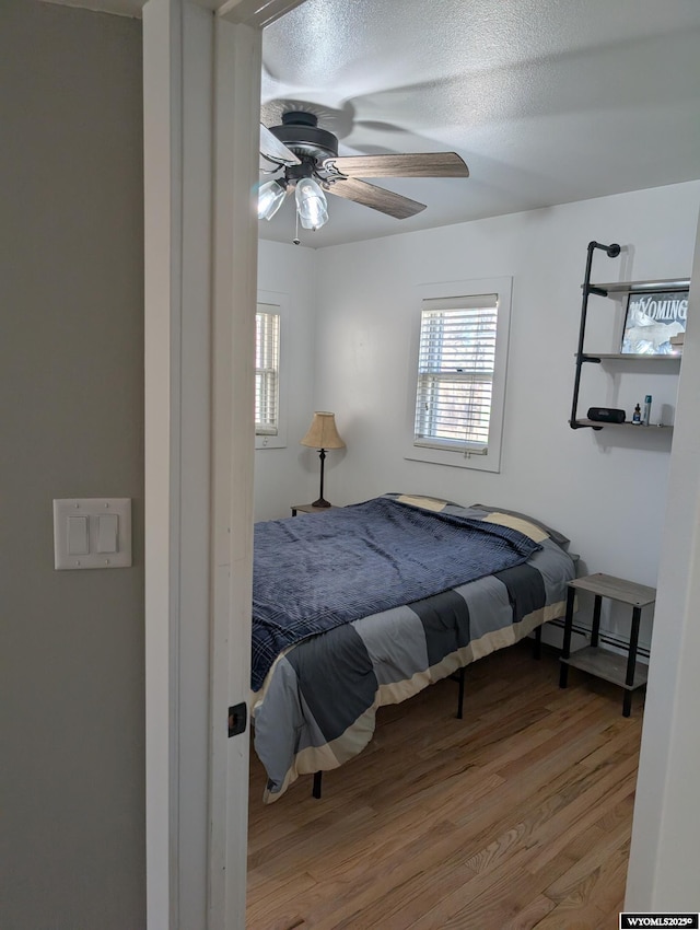 bedroom with a textured ceiling, a baseboard heating unit, wood finished floors, and a ceiling fan