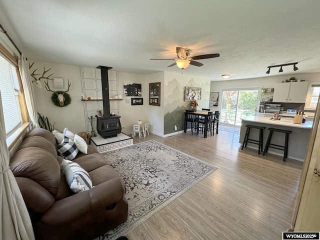 living area featuring baseboards, a wood stove, ceiling fan, light wood-style floors, and a textured ceiling