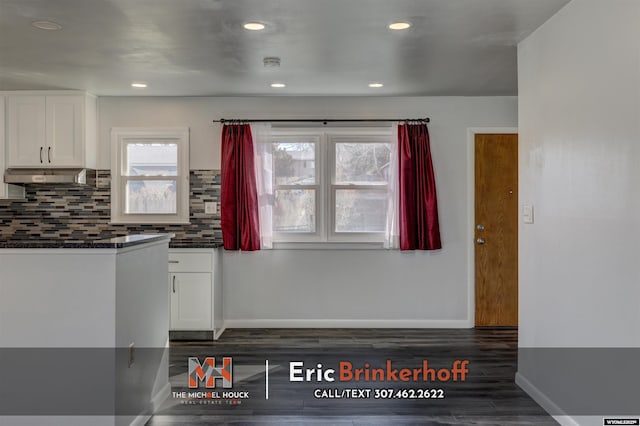 kitchen with dark wood finished floors, under cabinet range hood, white cabinetry, dark countertops, and tasteful backsplash