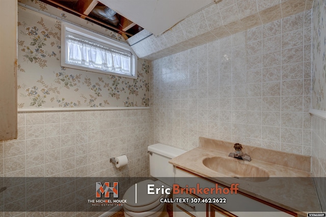 half bathroom featuring vanity, wallpapered walls, wainscoting, tile walls, and toilet