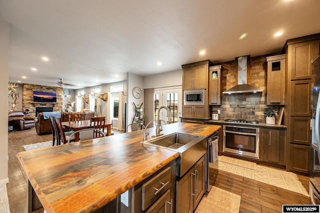 kitchen with a stone fireplace, stainless steel appliances, wall chimney range hood, wood counters, and decorative backsplash
