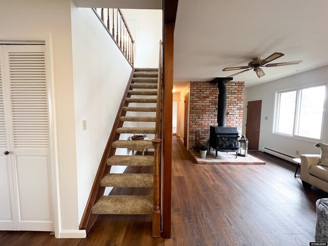 stairway featuring ceiling fan, a wood stove, wood finished floors, baseboards, and a baseboard heating unit