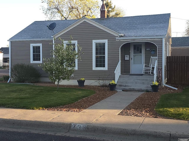 view of front facade with covered porch, a chimney, a front yard, roof with shingles, and fence