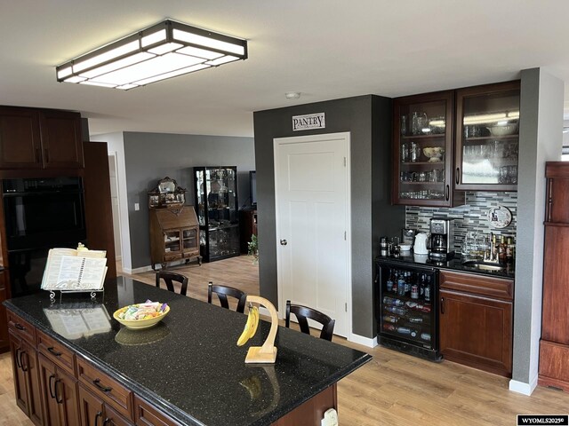 kitchen featuring beverage cooler, light wood finished floors, glass insert cabinets, dark stone counters, and decorative backsplash