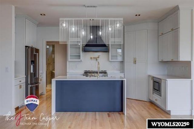 kitchen with stainless steel fridge with ice dispenser, built in microwave, light countertops, light wood-style floors, and a kitchen island with sink