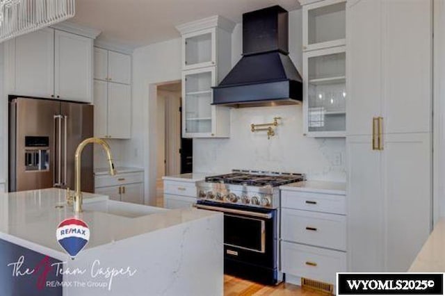 kitchen featuring stainless steel appliances, premium range hood, white cabinets, a sink, and an island with sink