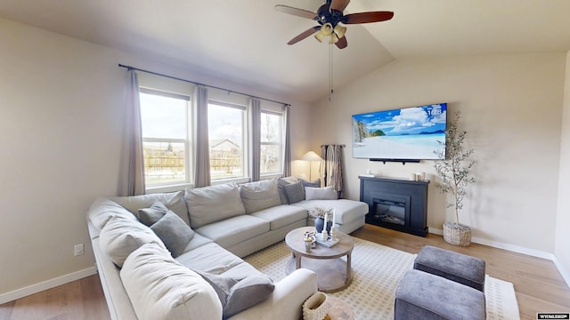 living area featuring a ceiling fan, lofted ceiling, light wood-style floors, a glass covered fireplace, and baseboards