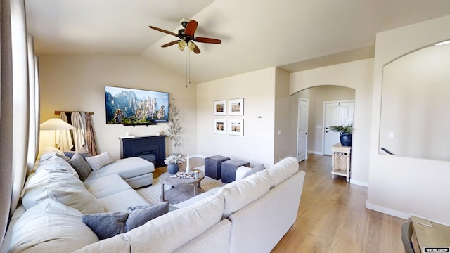 living room with a ceiling fan, arched walkways, light wood-type flooring, vaulted ceiling, and baseboards