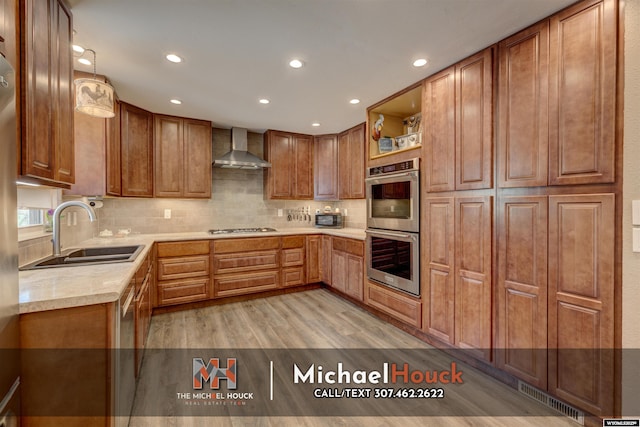 kitchen with a sink, wall chimney range hood, stainless steel appliances, light wood-style floors, and brown cabinetry