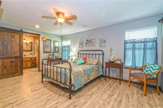 bedroom with a barn door, light wood-type flooring, connected bathroom, and ceiling fan