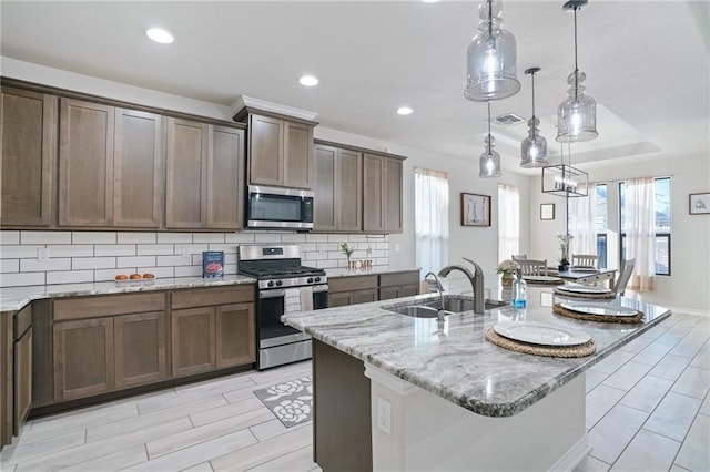 kitchen with light stone counters, stainless steel appliances, sink, decorative light fixtures, and a center island with sink