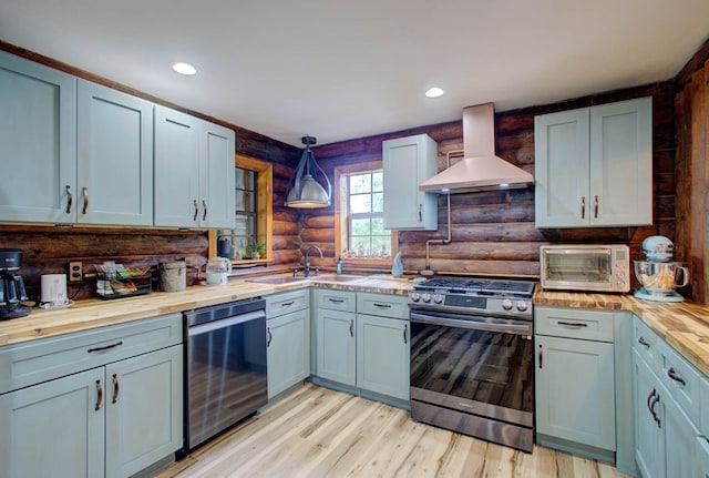 kitchen with pendant lighting, sink, butcher block counters, stainless steel appliances, and wall chimney exhaust hood