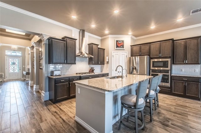 kitchen with a sink, wood finished floors, stainless steel appliances, wall chimney range hood, and ornate columns